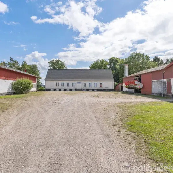 Et hvidt hus står centralt omgivet af røde ladebygninger under en skyet, men solrig himmel. Foran huset ligger en grusvej, mens træer omkranser scenen i baggrunden.