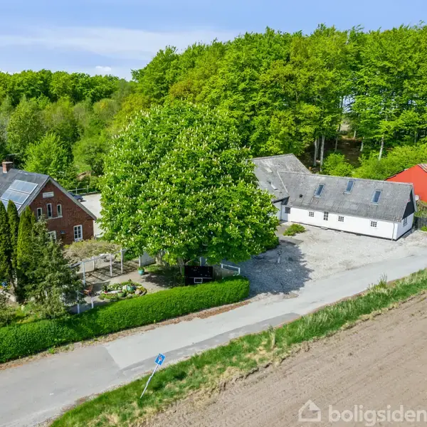 En gård med flere bygninger, herunder en rød lade og et hvidt hus. Gården er omgivet af træer og er placeret ved en vej med marker i forgrunden.