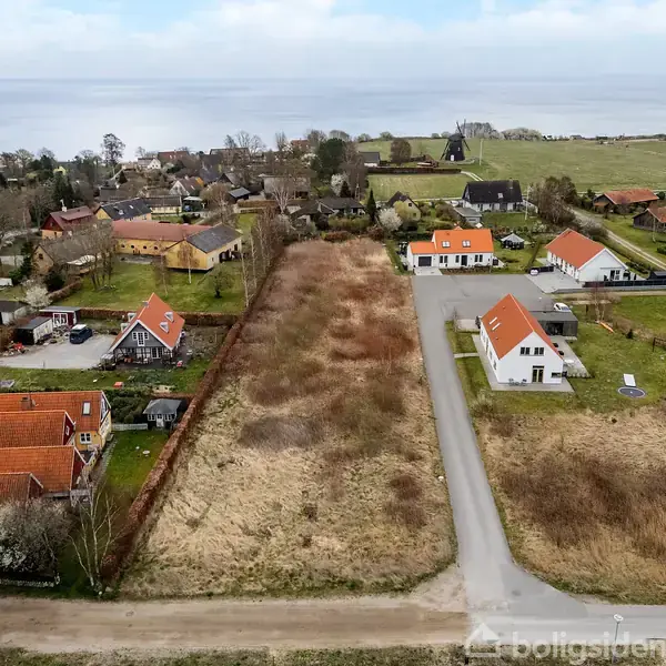 Et stort tomt område med højt græs omgivet af flere huse med røde tegltage i en landlig landsby med marker i baggrunden. En vej løber langs højre side af grunden.