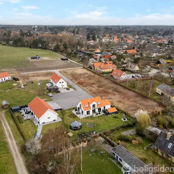 Et parcelhuskvarter med huse med røde tage, omgivet af grønne områder og marker. Haverne er velplejede, og en grusvej løber gennem nabolaget, der er stille og landligt.