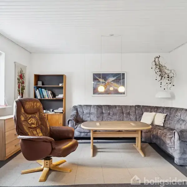 Brun lænestol står foran en grå hjørnesofa med et ovalt træbord imellem. Rummet er lyst med hvidmalede vægge, et vindue til venstre, reol og kunstværker.
