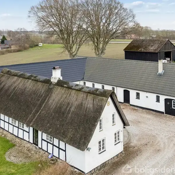 Hvidkalket gård med stråtag og teglsten, ligger i landligt område med grønne marker. Gården er L-formet, omgivet af grusvej og omkranset af store løvtræer og en rød traktor.
