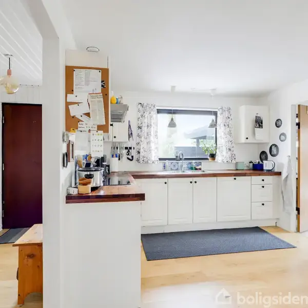 Et lyst køkken med hvide skabe og sorte greb, hvor der er en bordplade ved et stort vindue. Kontekst: Entré til venstre med sko, trædør og farverig måtte.