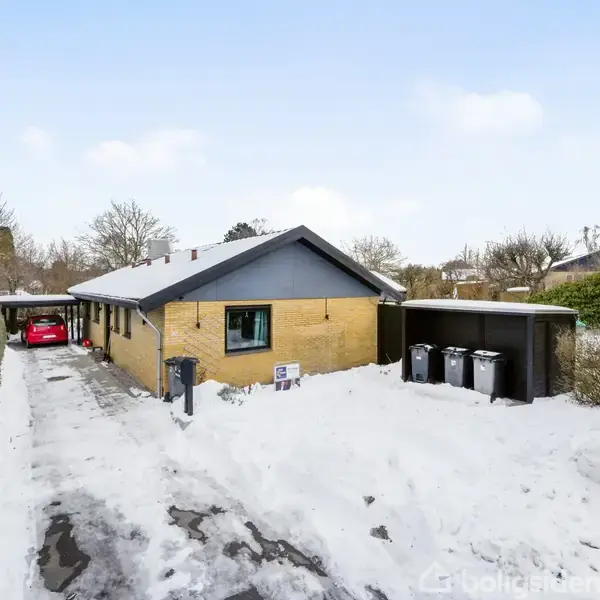 Et enkeltstående hus af mursten står i en snedækket have. Tre skraldespande er placeret udendørs. En rød bil er parkeret under en carport til venstre ved siden af huset.
