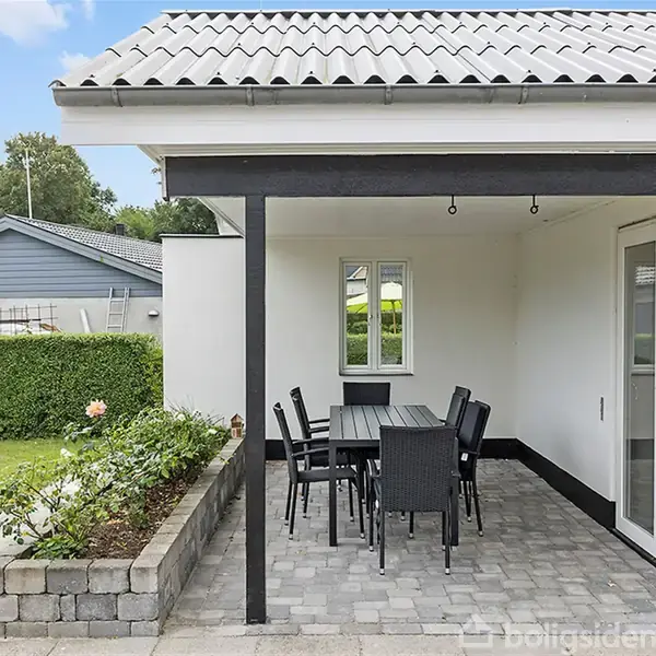 Et havebord med seks stole står under en sort overdækket terrasse ved et hvidt hus. Terrassen er omgivet af grønne buske og en græsplæne.