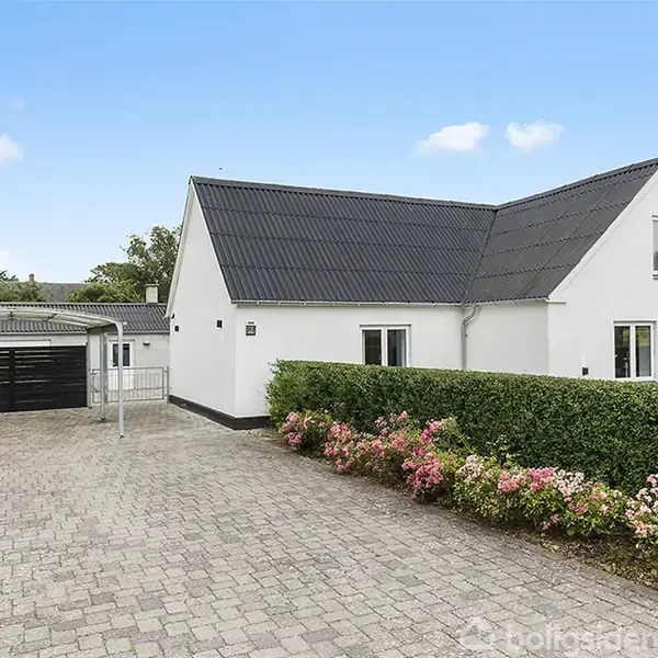 Hvidt hus står stille ved siden af brolagt indkørsel, flankeret af hæk og blomster, i et forstadsområde med spredte træer i baggrunden.