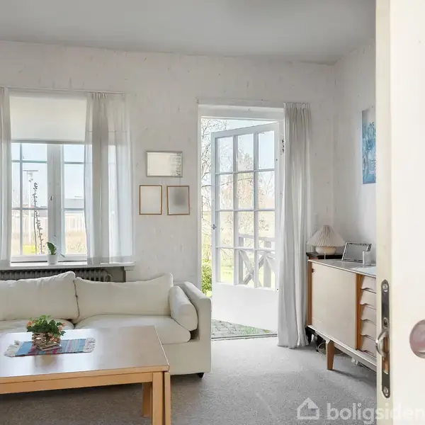 Hvid sofa står ved et træbord i stue med lyse vægge. Lige ved vinduet fører en glasdør ud til en terrasse. Enkel indretning med lysindfald fra vinduet.