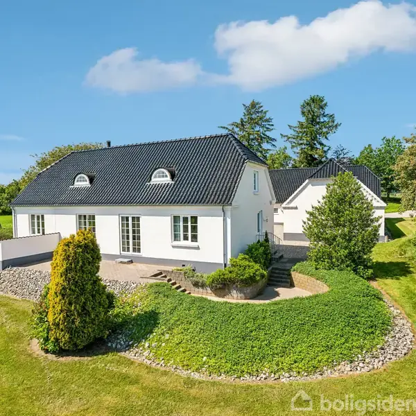 Hvidt hus med sort tag står omgivet af grøn have og træer; beliggende i landligt område under blå himmel med få skyer.