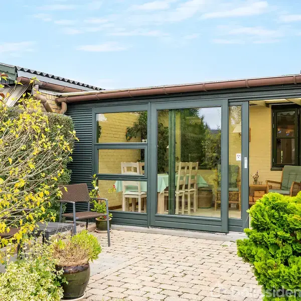 Terrassedør af glas åbner ind til en lys stue med spisebord. Omkranset af gul murstensvægge, grønne buske og en brostensbelagt terrasse i et boligområde.