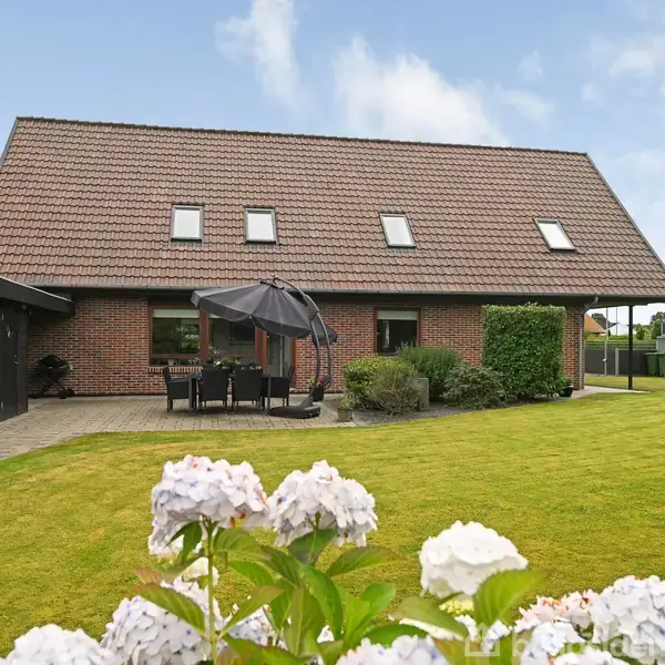Et rødt murstenshus med en stejl taghældning og flere små vinduer. Foran huset er en terrasse med et bord og parasol. Haven har en velplejet græsplæne og blomster.