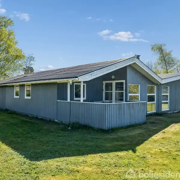 Et blåmalet træhus står i en grøn have med velholdt græs. Huset har store vinduer og et skråt tag. Bagerst ses træer og en klar himmel med få skyer.