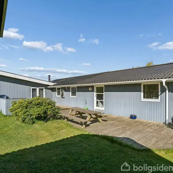 En blåmalet træbygning står stille, omgivet af velplejede buske og græs, med en træbænk på en terrasse under en klar blå himmel.