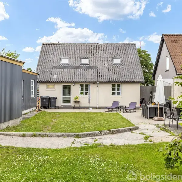 Hus med gråt tag og hvide vægge står centralt. To liggestole er foran huset. Til venstre er en skurbygning, til højre en terrasse med parasol og havemøbler. Græsplæne omgiver.