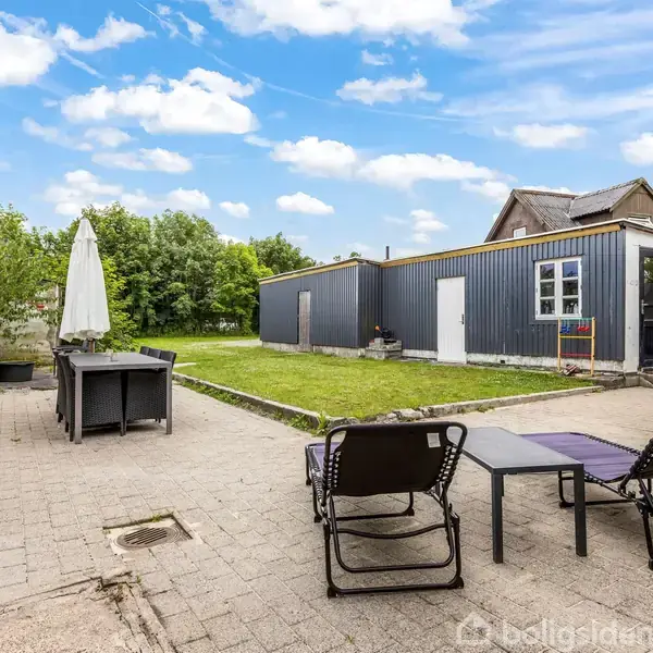 Terrassemøbler står på en flisebelagt terrasse; en parasol er åben ved siden af et bord. I baggrunden er der en græsplæne og et hus med gråt ydre. Skyscenen er klar.