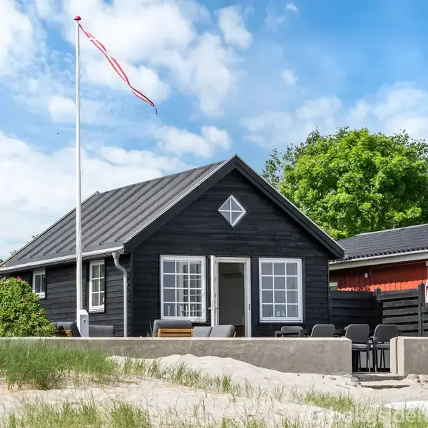 Sort sommerhus med hvide vinduer og terrasse står på en sandet grund. Et flag hejses i flagstangen ved siden af huset. Grønne buske og en rød bygning ses i baggrunden.