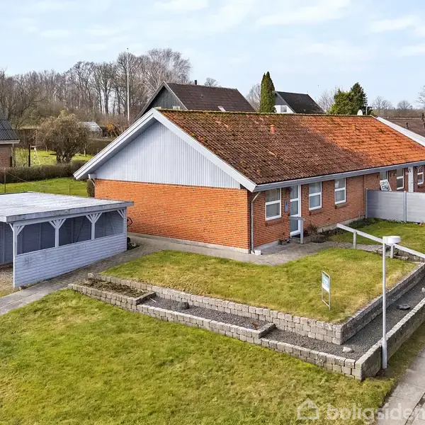 Rødt murstenshus med tag og vinduer, flankeret af en blå garage til venstre. Huset står på en hævet græsplæne ved en stille boligvej i et villakvarter.