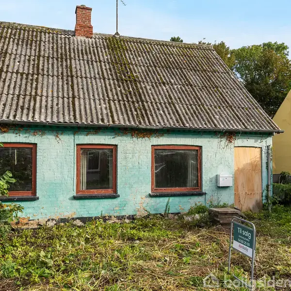 Et lille, ældre hus med gråbrunt bølgeeternittag står i en have med uklippet græs. Hoveddøren sidder til højre under en stor vinduesramme. Skilt foran: "Til salg".