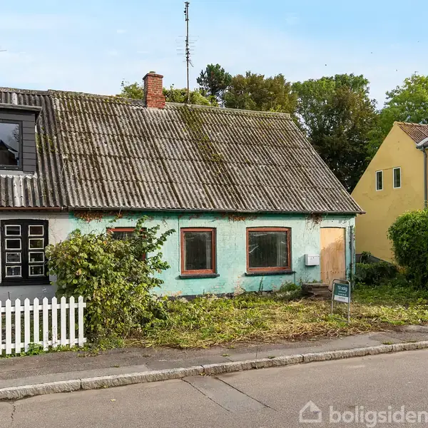 Et ældre, slidt hus står stille med en blå facade og gammelt tag. Ved siden af huset er der en gul bygning og en hvid hegnssektion. En vej løber foran.