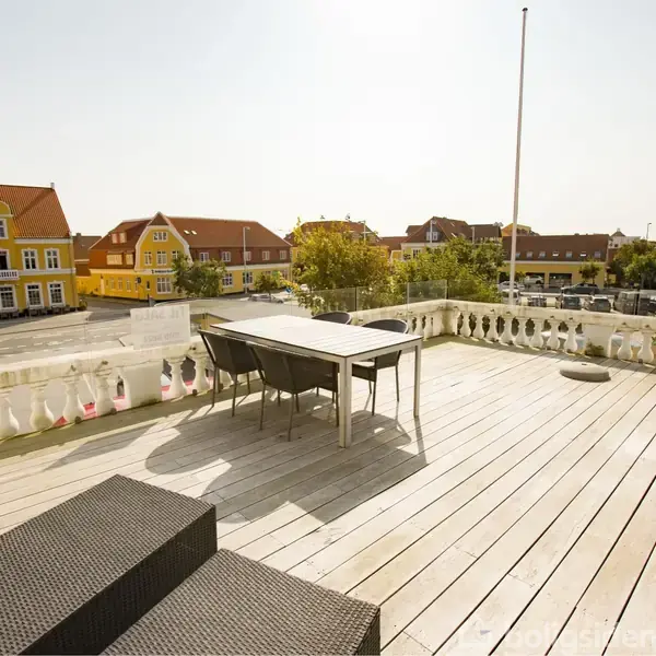 Et bord med stole står på en stor terrasse med trægulv. I baggrunden ses farverige bygninger under en klar himmel.