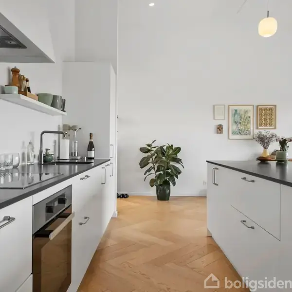 Køkken med hvide skabe og sorte bordplader. Et stort køkkenbord er centralt placeret. En plante står i baggrunden. Væggene er dekoreret med kunstværker, og en TV-skærm er synlig.