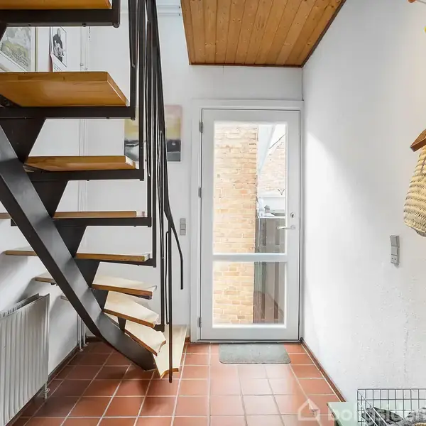 Trappe med trætrin fører opad; rummet har hvide vægge og rødbrune fliser. En glasdør viser en murstensvæg udenfor. En radiators placering er under trappen.