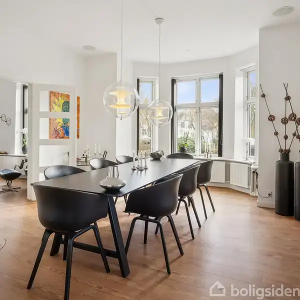 Spisebord med sorte stole står under tre moderne lamper. Væggene prydes af farverige malerier. Store vinduer bagved giver lys til rummet, som har trægulve og en åben indretningsstil.