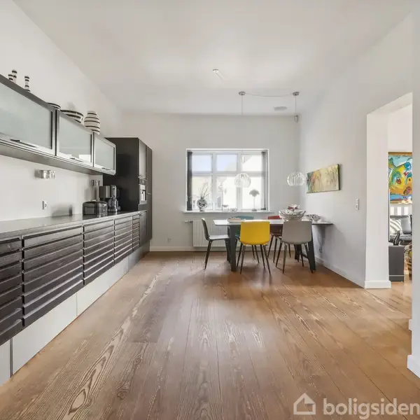 Køkken med lange, mørke skabe langs venstre væg, flere dekorative vaser ovenpå, og et spisebord med forskellige stole mod vinduet. Rummet har lyse træplanker på gulvet.