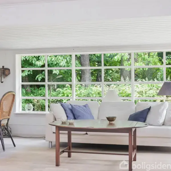 Sofa med blå puder står foran et stort vindue med udsigt til en grøn have. Rundt sofabord i forgrunden, stole ved spisebord til venstre, lampe til højre.
