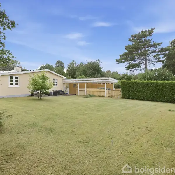 Et lille gult hus står i en velplejet have med græsplæne og træer omkring. Der ses også en carport ved siden af huset. Himlen er blå med få skyer.