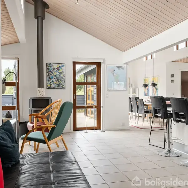 Sofa med farverige puder står i stue med flisegulv; en brændeovn ved siden af. Spisebord og barstole i det åbne køkkenområde, med lysindfald fra vinduer og dør.