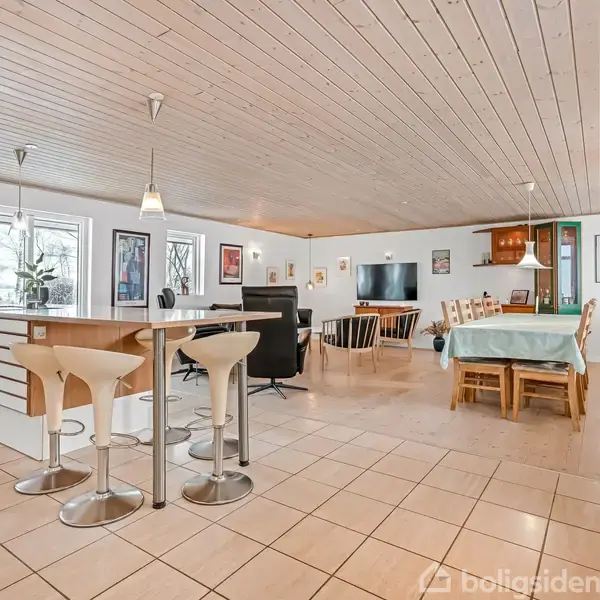 Et åbent køkkenområde med en bar og to høje stole. I baggrunden er en stue med spisebord og tv, omgivet af lyse vægge og flisegulv. Ingen tekst.