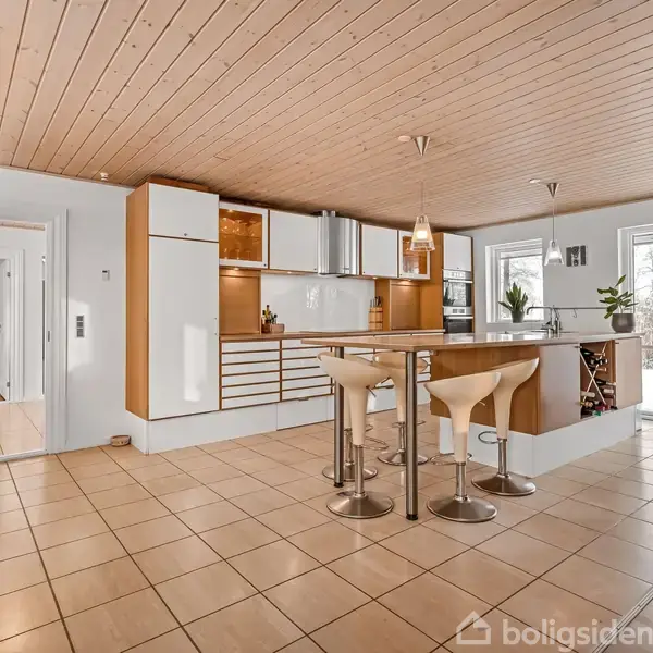 Køkken med moderne design, køkkenø med barstole og højt til loftet, omgivet af skabe og apparater. Store vinduer til venstre bringer naturligt lys ind.