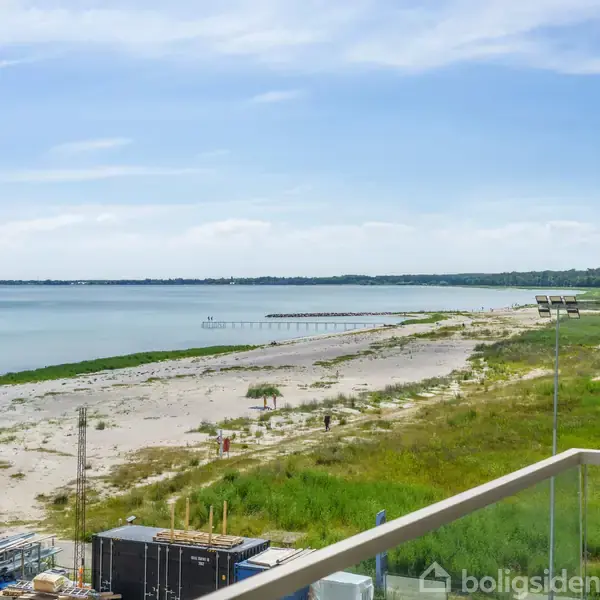 Strand med sand og vegetation strækker sig langs en kystlinje. En mole fører ud i vandet. I forgrunden ses en glasbalkon, mens himlen er blå med få skyer.