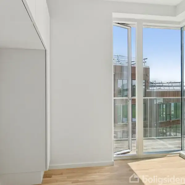 Et lyst værelse med parketgulv har store vinduer, der åbner mod en balkon med udsigt over moderne bygninger og vandet udenfor.
