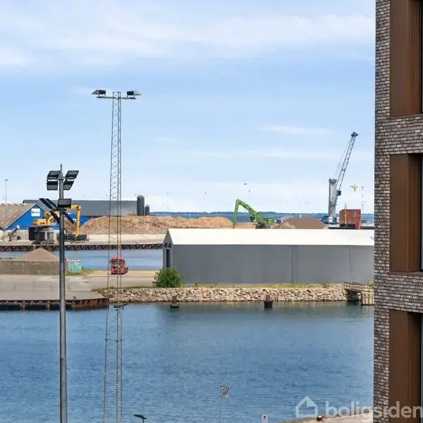 Bygning står stille ved havn, med kraner i baggrunden og en blå himmel over vandet. En moderne lejlighedskompleks er til højre, mens industriområde kan ses til venstre.