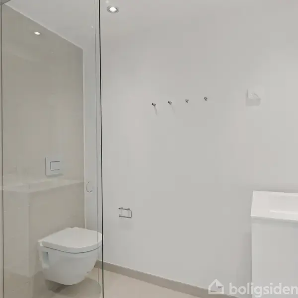 Badeværelse med væghængt toilet og hvid vask. Bruseren med glasvæg ses til venstre. Rummet er minimalistisk indrettet med hvide vægge, beige gulv og spotlight-belysning.