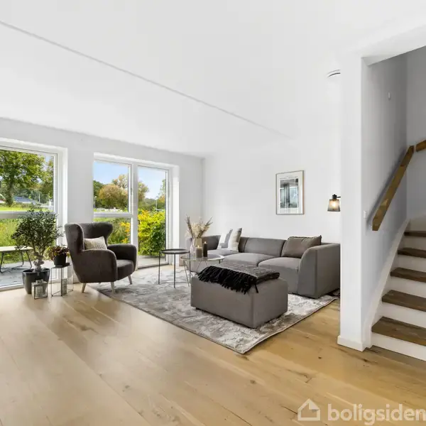 Stue med grå sofa og lænestole på tæppe, store vinduer med udsigt til grøn have. Trætrappe fører til øvre etage. Pejseplante og dekorative elementer skaber hyggelig atmosfære.