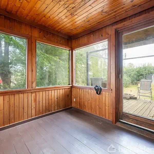 Et rum med trævægge og trægulv har store vinduer, giver udsigt til en grøn have med træer og en terrasse. Lyset strømmer ind, skaber en naturlig atmosfære.