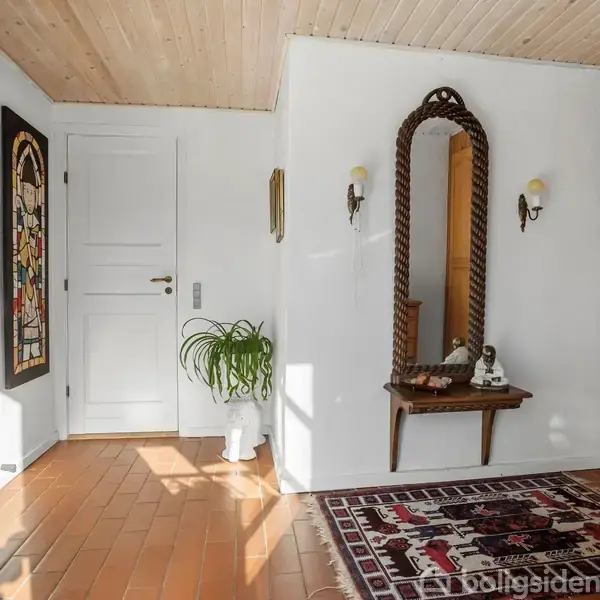 Et indgangsparti med et stort vægspejl og et bord midtfor. Til højre er der et træskab, og en plante står ved en hvid dør. Sollys strømmer ind.
