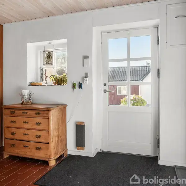 En trækommode står ved en hvid væg med en glasdør. En lille vindueskarm indeholder planter og dekorationer. Rummet har en bøjlestang og flisegulv. Udsigt til et rødt hus udenfor.