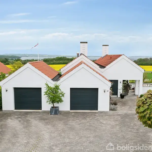 Et hvidt hus med tre sorte garageporte står på en brostensbelagt gårdsplads. Omkring huset er der grønne hække og træer, med marker og hav i baggrunden.