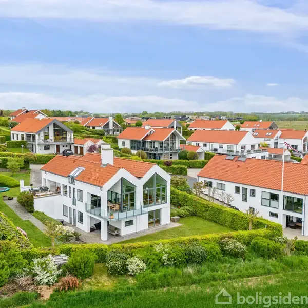 Huse med røde tage og hvide facader står stille i en grøn og frodig villakvarter. Blå himmel og grønne arealer omgiver området, som består af flere lignende boliger.