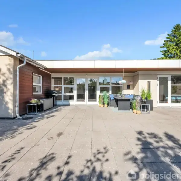 Moderne hus med store vinduer og terrassedøre åbner ud mod en rummelig fliseterrasse. Terrassen har udendørs møbler og planter i krukker. Der ses en klar blå himmel i baggrunden.