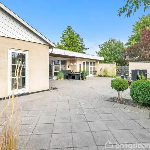 Hus med fladt tag og store vinduer. Terrasse med fliser, dekoreret med planter i krukker. Træer og buske omgiver området, skaber en hyggelig, privat atmosfære.