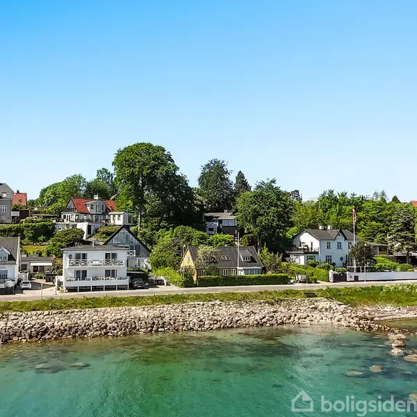 En række huse står langs en kystvej med havet på den ene side og træer samt bygninger på den anden. En bil kører på vejen.