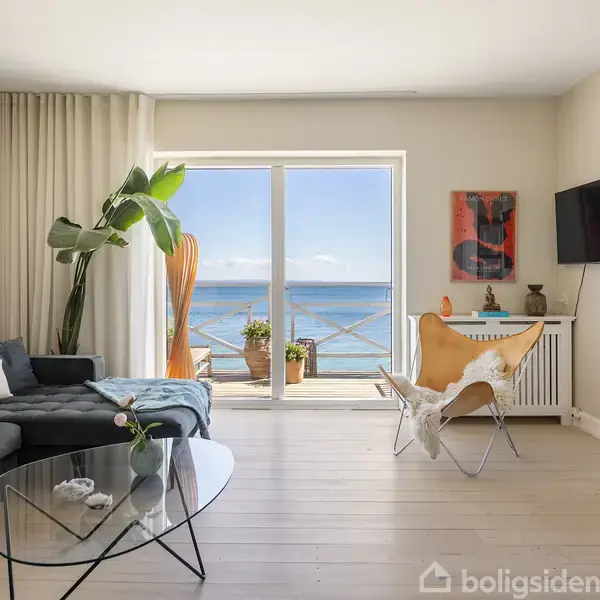 Sort sofa i en stue står overfor et glasbord, med en lænestol ved siden af. Store vinduer viser havudsigt. En plante og kunstværker pynter rummet. Tekst: "boligsiden".