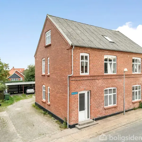 Rødt murstenshus står stille i et beboelsesområde med andre huse. Ved siden af huset er en grusvej, der fører til en carport og en have i baggrunden.