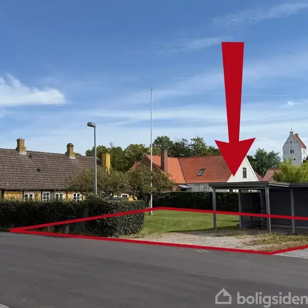 Rød pil og omrids markerer en parcel med carport og hæk ved en landevej. Flere gule og røde huse samt en kirke ses i baggrunden under en blå himmel.