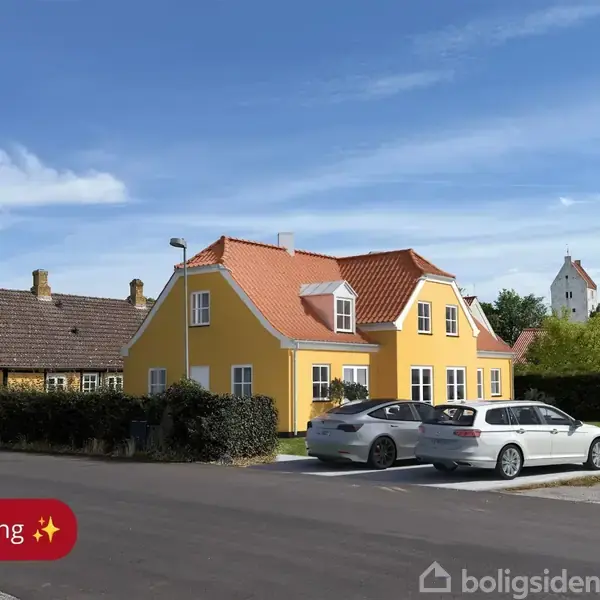 Gult hus med rødt tag står stille ved vej, omgivet af grønt hegn og to parkerede biler. Et tårn ses i baggrunden. Tekst: "AI-visualisering ✨".