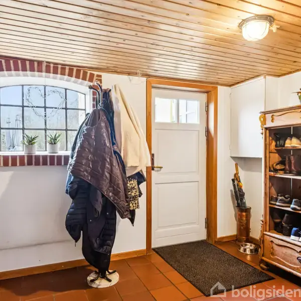 Entré med tøjstativ fyldt med jakker, en skohylde til højre med sko og en dør i midten. Et vindue med murstensbue ovenfor tilføjer naturligt lys. Træloft og flisegulv.
